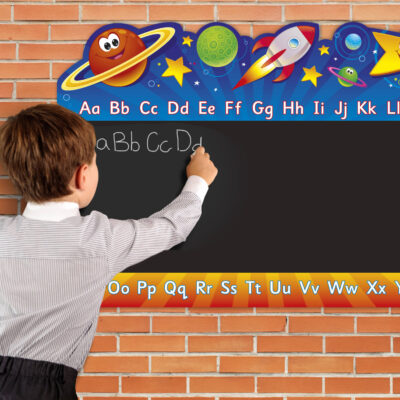 Male child to left of image, writing onto sign. The sign is colourful space themed illustrations. Including planets with friendly faces. English alphabet A to Z, top and bottom of black chalkboard that is within the central area of the sign.